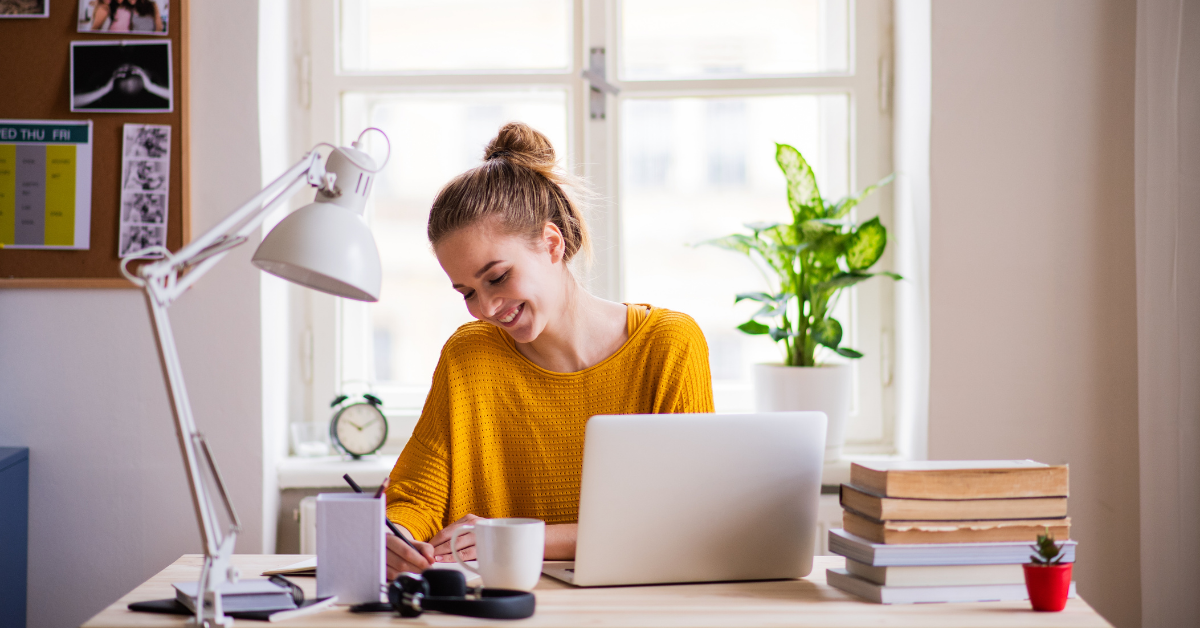 A woman in a yellow shirt smiles while working at a desk from home with office items around her.