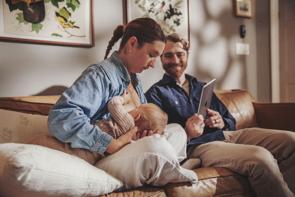 A woman breastfeeds her baby while her partner holds an iPad for a telehealth lactation consultation.