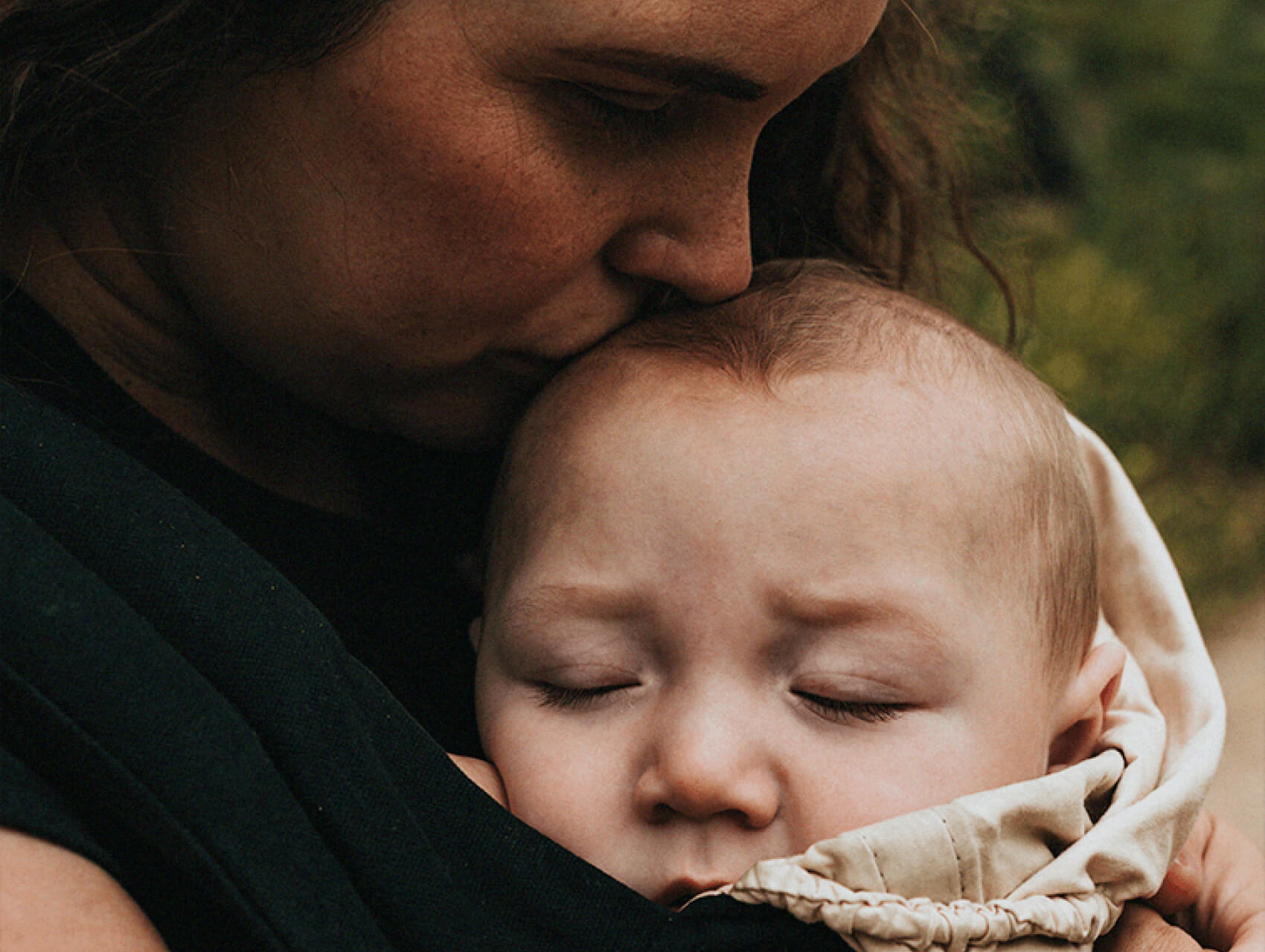 A parent kissing their baby’s head while carrying them in a wrap for a cozy nap.