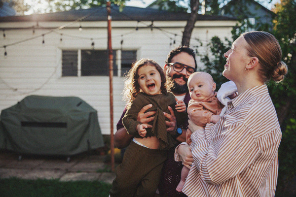 two parents hold infant and small child in backyard while smiling
