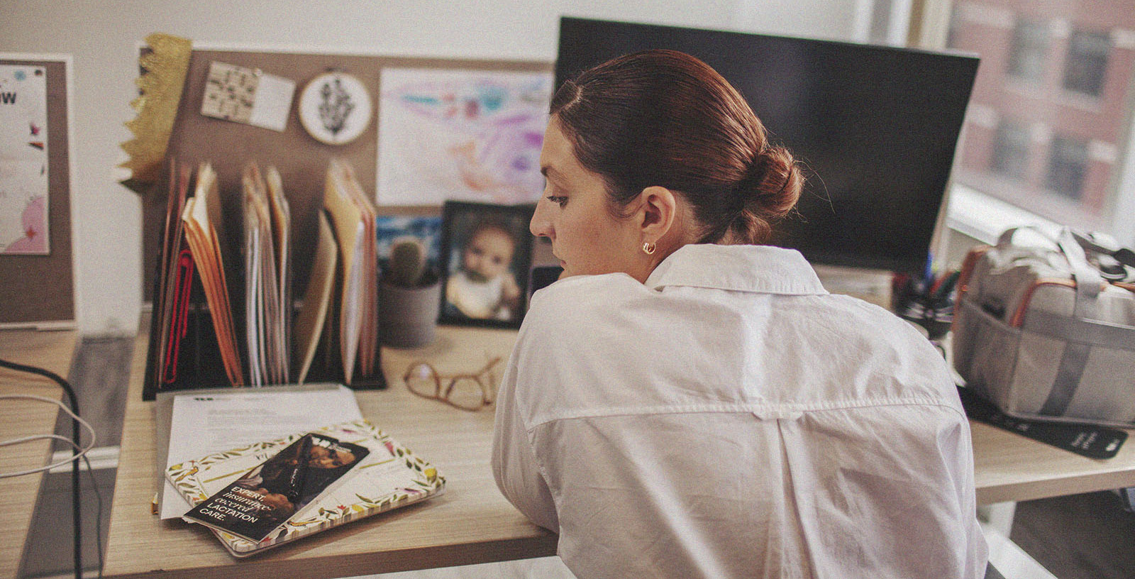 A woman sits at her office desk, facing away from the camera. She looks over at a lactation care brochure on the edge of the desk.