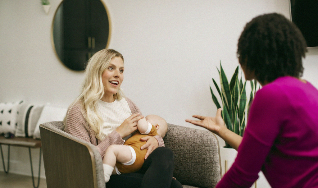 A smiling woman of European descent sits in a comfortable chair and nurses her infant while speaking with a woman of African descent.