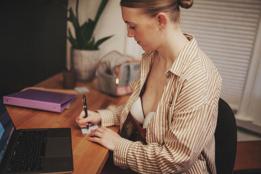 A woman sits at a desk while writing. She wears a collared shirt and her breast pump is visible.