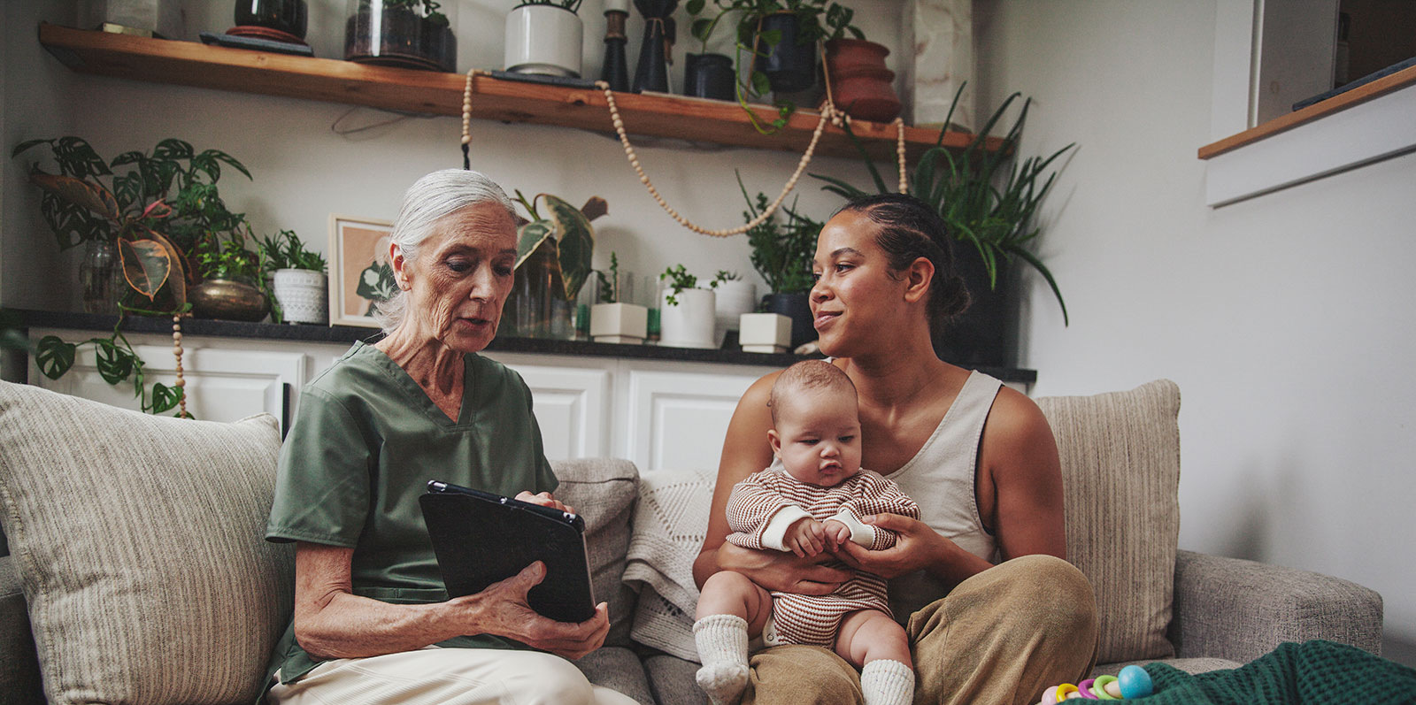 An IBCLC points to items on a tablet while seated on a sofa next to a mom and her infant during a home visits