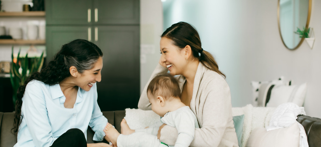 A mother laughs while interacting with a smiling IBCLC, who is playing with her baby on her lap.