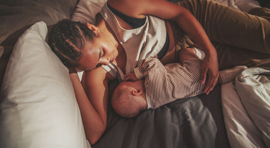 Woman cradles and breastfeeds baby while gazing gently at their face and lying on her side in bed.