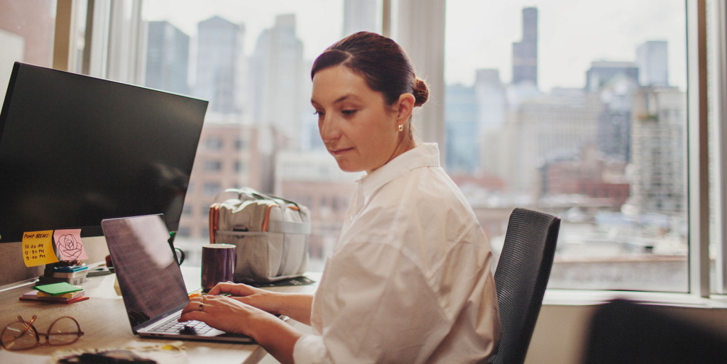 A woman sits at her desk at work with her breast pump bag and a pump break schedule within view.