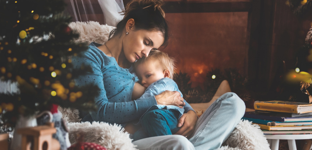 A parent nestles a young, breastfeeding baby in their arms while sitting on the ground in front of a sparkling Christmas tree.