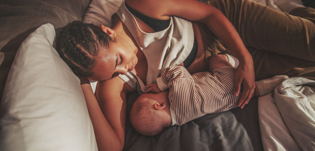 A woman of African descent lays on her couch in the afternoon, gazing at her young baby while she breastfeeds him