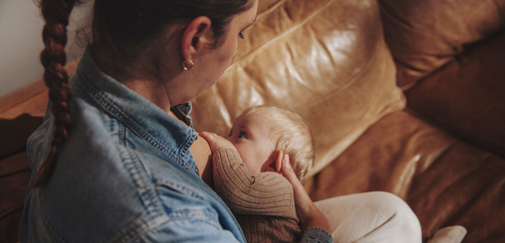 A parent with long brown hair in a denim shirt looks down on her baby while they breastfeed on a sofa.