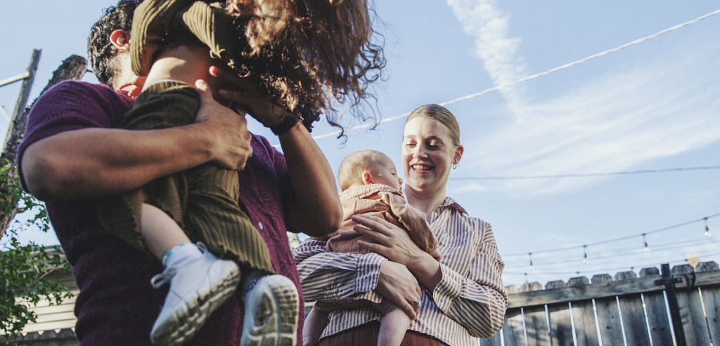 Two parents play with their two young children in a yard, swinging them around and laughing.