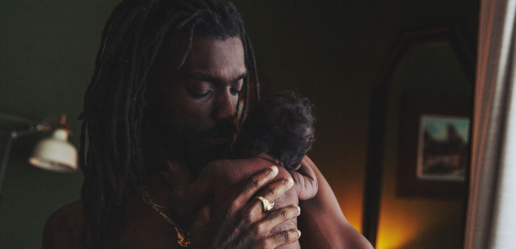 A father with dreadlocks cradles a newborn in a softly lit room while his eyes are closed.